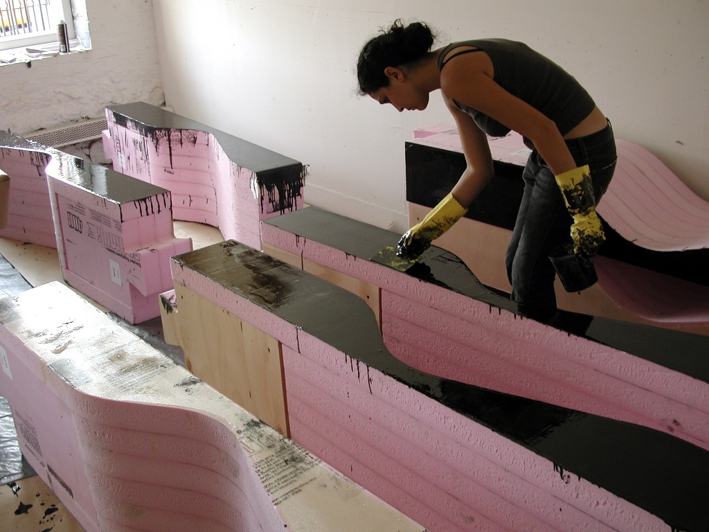 A worker coats one of the foam and wood laminated chaise lounges with epoxy tinted with graphite powder.